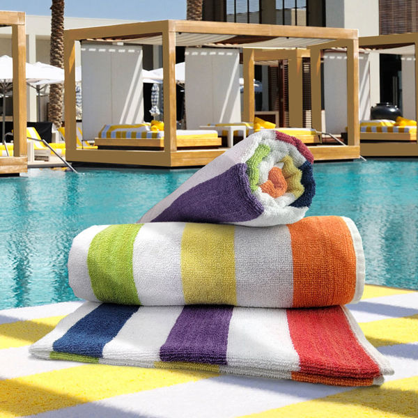 Two Ganesh Mills Promotional Beach Towels - 2x2 Cabana Multicolor Stripe are stacked on a lounge chair by the pool, with cabanas and palm trees in the sunny background.