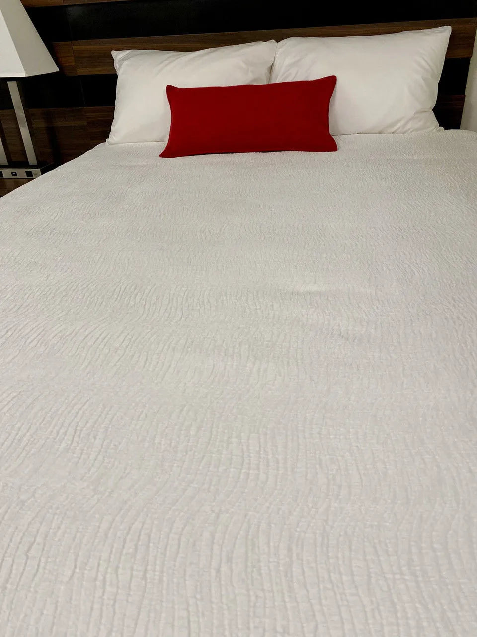 A queen and king bed topped with the Ganesh Mills Bed Topper in a jacquard wave design, two white pillows, and a red accent pillow in front. A lamp sits on the nightstand to the left.