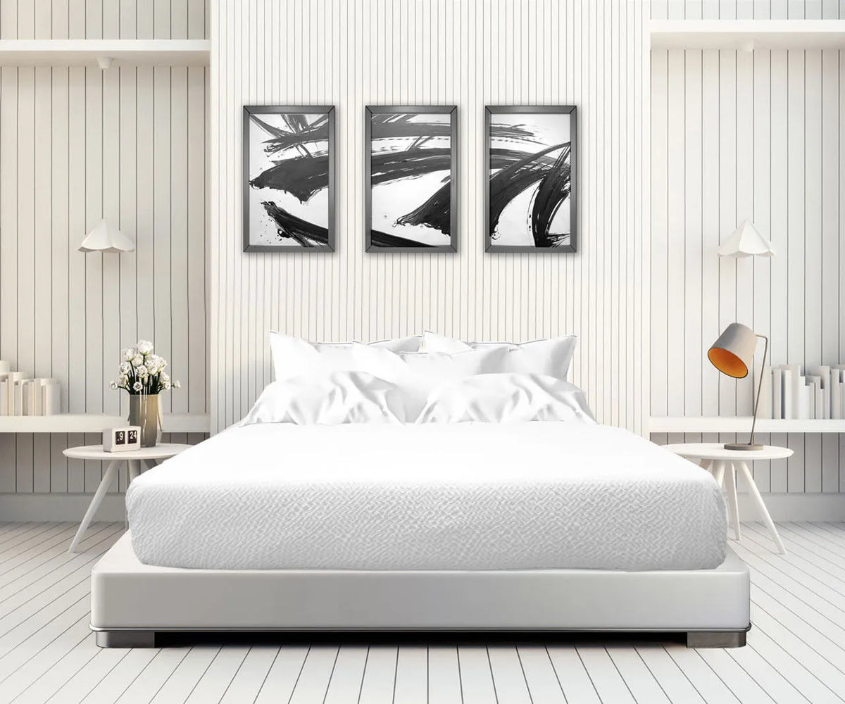 A minimalist bedroom features a white bed with the Ganesh Mills Oxford Bed Topper - Crinkle Weave, side tables, modern lamps, and three abstract black-and-white paintings above. White walls, shelves with books, and a vase of flowers complete the look.