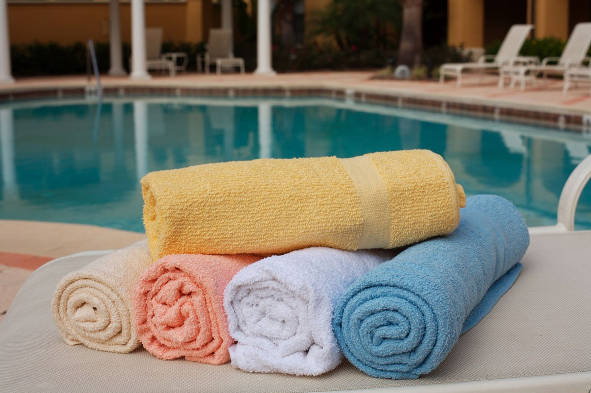 Four Oxford Solid Color Pool Towels (Bronze-Classic) by Ganesh Mills in yellow, orange, white, and blue are stacked on a lounge chair by the pool with patio furniture nearby—ideal hotel towels for relaxing by the water.