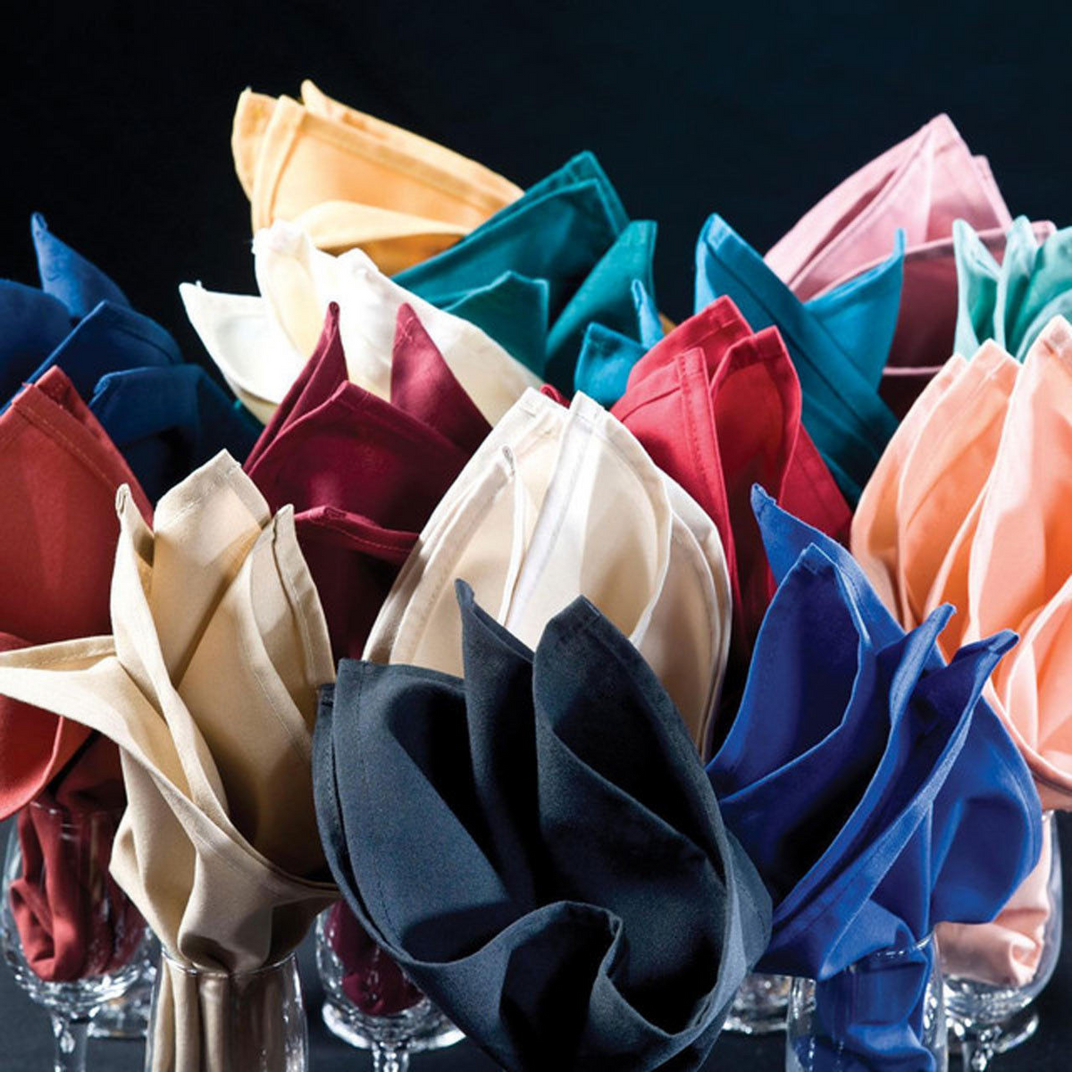 A collection of 1Concier Napkin Premier 100% poly Bermuda Sand napkins is elegantly folded and displayed upright in glasses, set closely together against a dark backdrop.