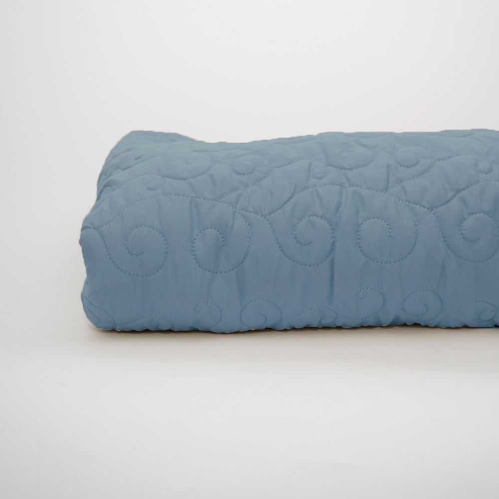 The Ascent Standard Bedspread by Calderon Textiles, featuring a folded light blue quilted design with swirling stitching, is displayed on a plain white background.