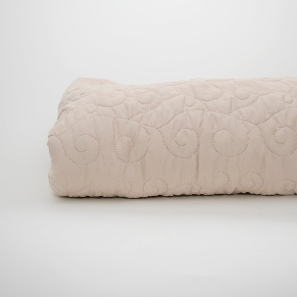 The Ascent Standard Bedspread by Calderon Textiles, featuring a beige quilted design with a subtle stitched swirl pattern, is folded and displayed on a plain white background.