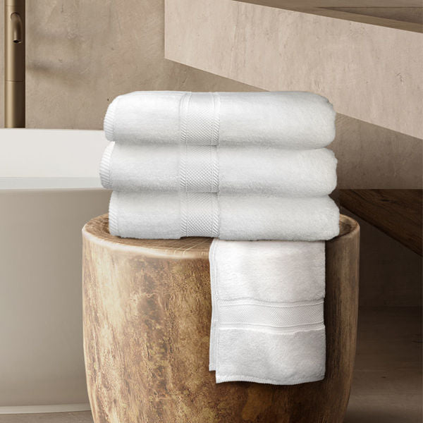 Three white Bath Towels from Ganesh Mills' Oxford Miasma collection are neatly stacked on a round wooden stool, with one towel draped over the edge, highlighting their plush elegance in the modern, neutral-toned bathroom.