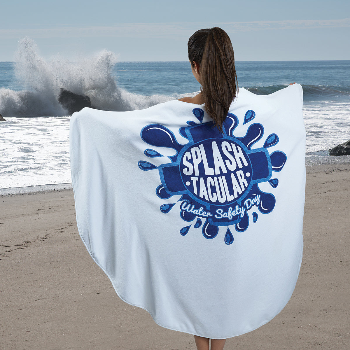 On a sandy beach, a person faces the ocean wrapped in a 60″ Round Beach Towel by Pro Towels featuring a blue SPLASH-TACULAR Water Safety Day logo, while waves crash under a partly cloudy sky.