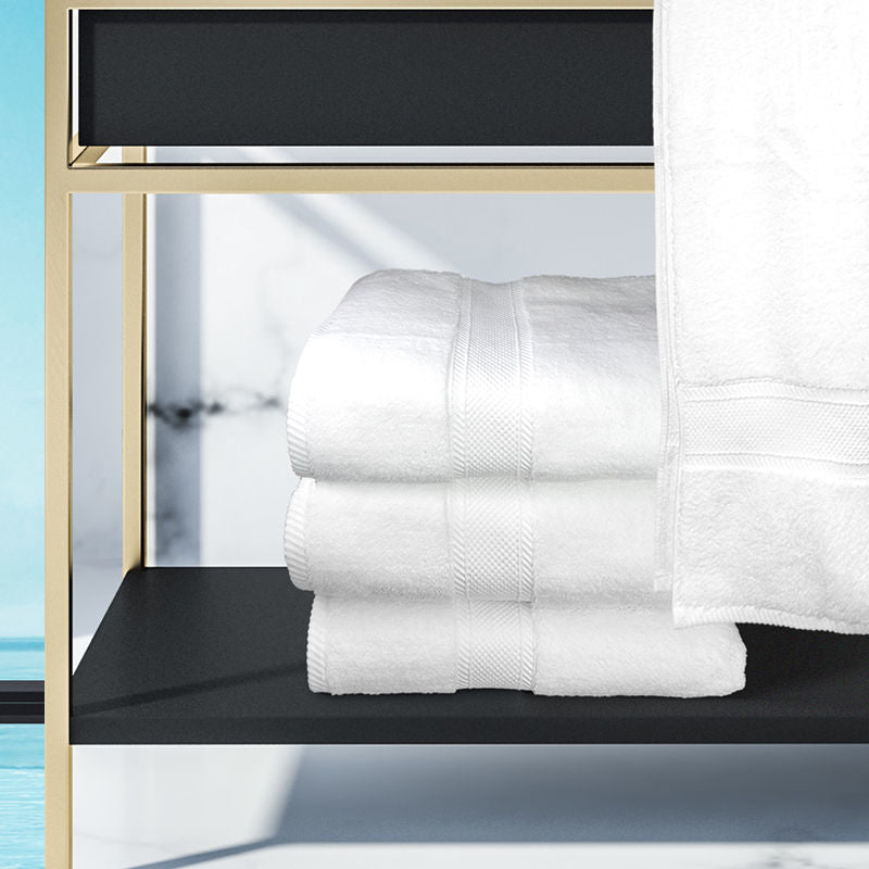 A white Bathmat from the Ganesh Mills Oxford Miasma Towel Collection is stacked on a black shelf with gold-colored framing, made from zero twist cotton for plush, hotel-like comfort. Bright light and a blue pool shine in the background.