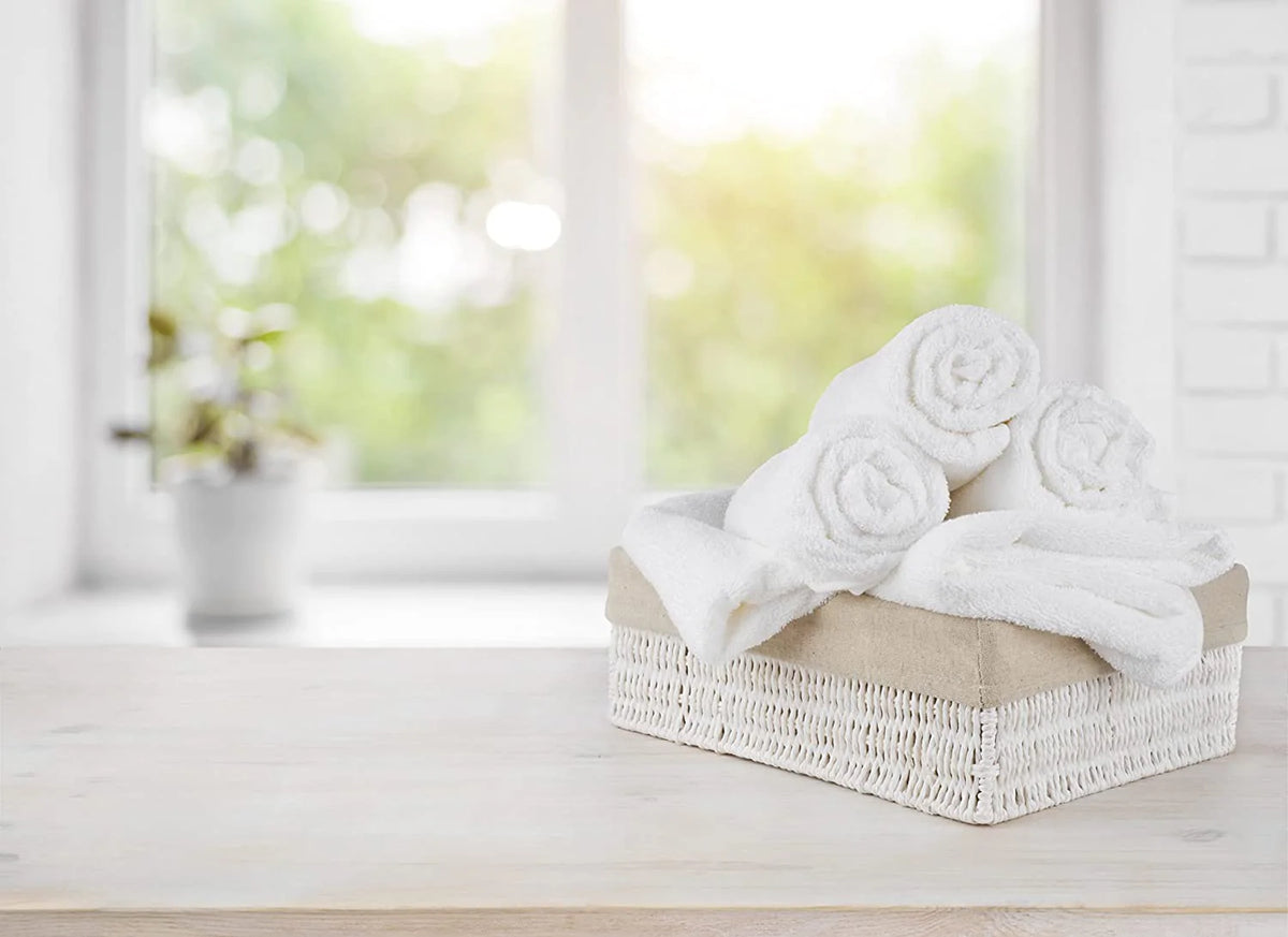 A wicker basket with neatly rolled KSE Bath Towel - Basic Economy 10 Single Towels sits on a wooden surface, with a blurred bright window and a potted plant in the background.