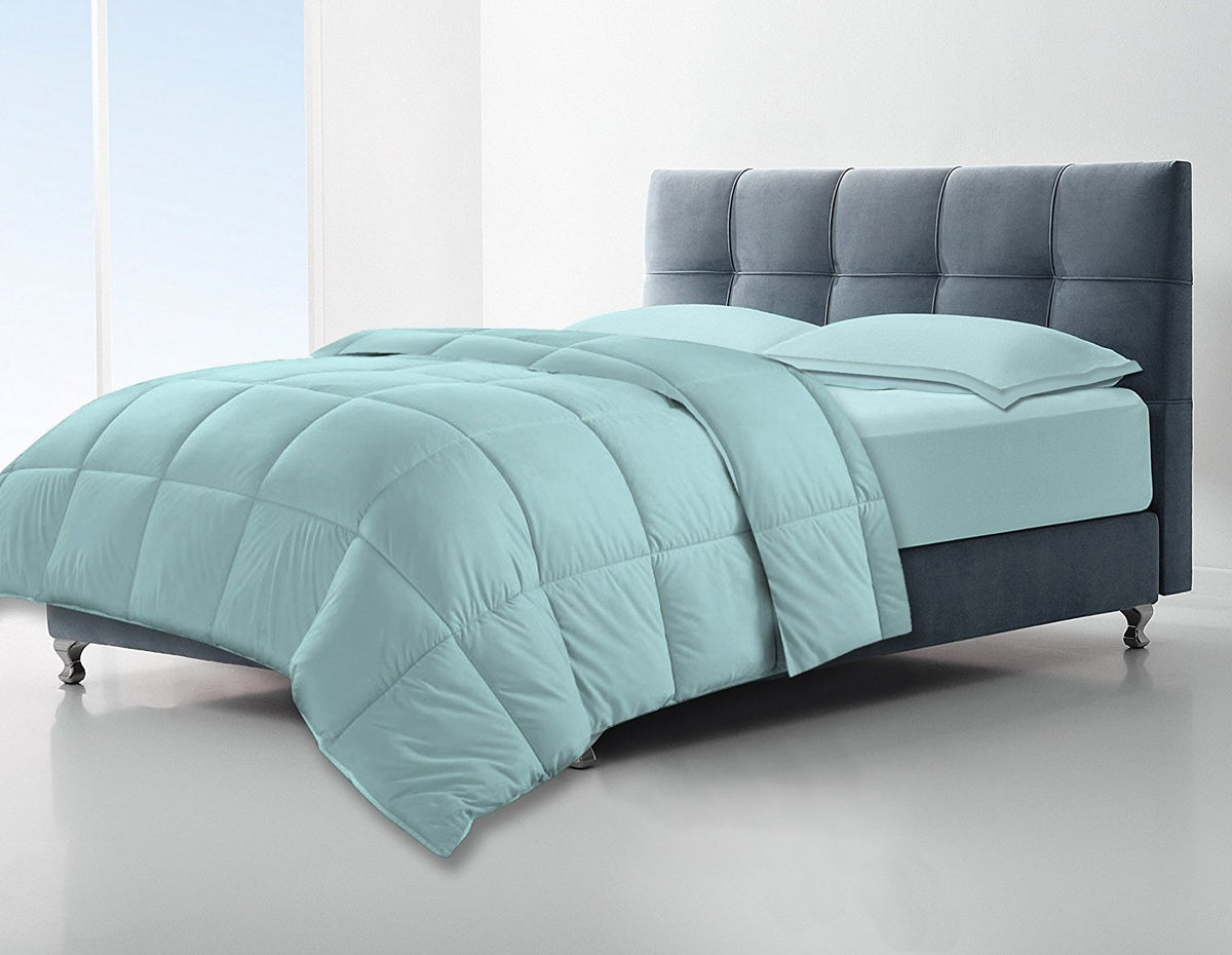 A modern, minimalistic bedroom features a neatly made bed with the KSE Down Alternative Comforter in light blue and matching pillows, set against a tufted dark gray headboard and pale walls, with natural light from large windows on the left.