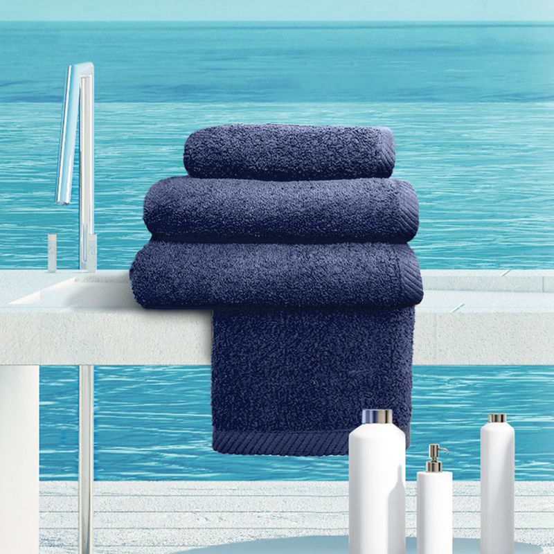 A stack of Ganesh Mills Bath Towel - Navy Blue Oxford Magic BleachGuard, made from ringspun cotton, is neatly folded on a white counter beside a sink, with white bottles and a tranquil blue ocean in the background.
