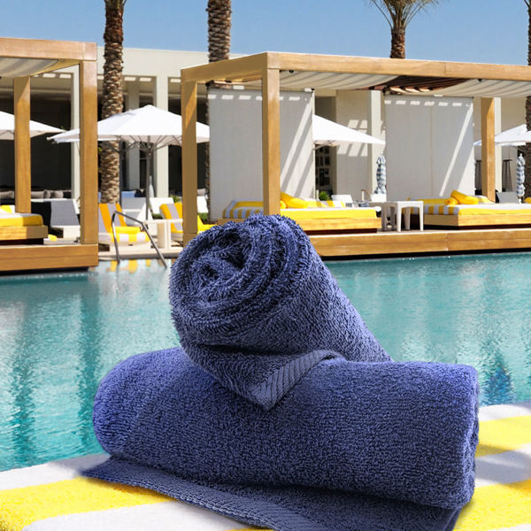 Two rolled Ganesh Mills Bath Towels in Navy Blue Oxford Magic BleachGuard are placed on a striped yellow and white surface by a pool, with cabanas, lounge chairs, and palm trees visible in the sunny background.