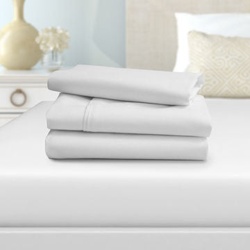 A neatly folded stack of Ganesh Mills Fitted Sheets - Oxford Super Deluxe Bed Linen rests on a made bed in a bright bedroom, with a nightstand, decorative vase, and pillows softly blurred in the background.