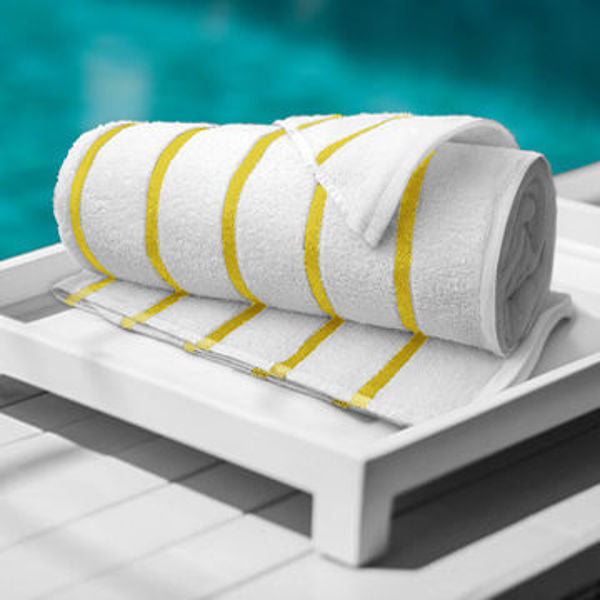 The Ganesh Mills Oxford Nautique Stripe Towel—White with Blue/Yellow Stripes—is rolled on a white tray by the pool, set atop a white wooden surface with the blurred blue water in the background.