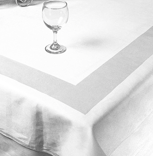 A single empty wine glass sits on a neatly laid white 1Concier Table Linen Cottonrich Ultimate, featuring a wide light satin band near the table’s edge.