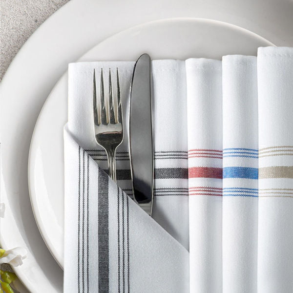 A fork and knife rest on a white plate atop a folded Ganesh Mills Bistro Napkin—Reverse Color with White Stripes. The plate also holds additional polyester napkins, each folded with red, blue, and tan stripes.