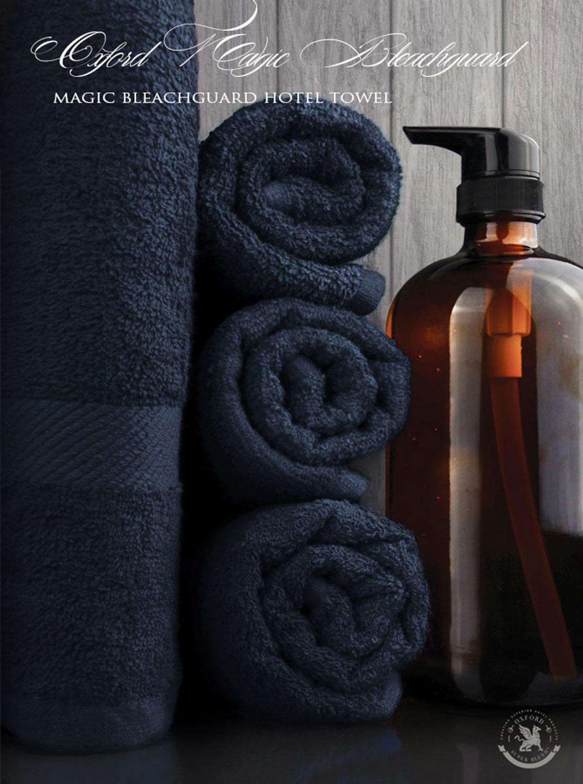 A stack of navy blue rolled towels and a folded towel sit beside an amber pump bottle against a dark background. Text reads: Ganesh Mills Bath Towel - Navy Blue Oxford Magic BleachGuard, perfect for bulk wholesale needs.