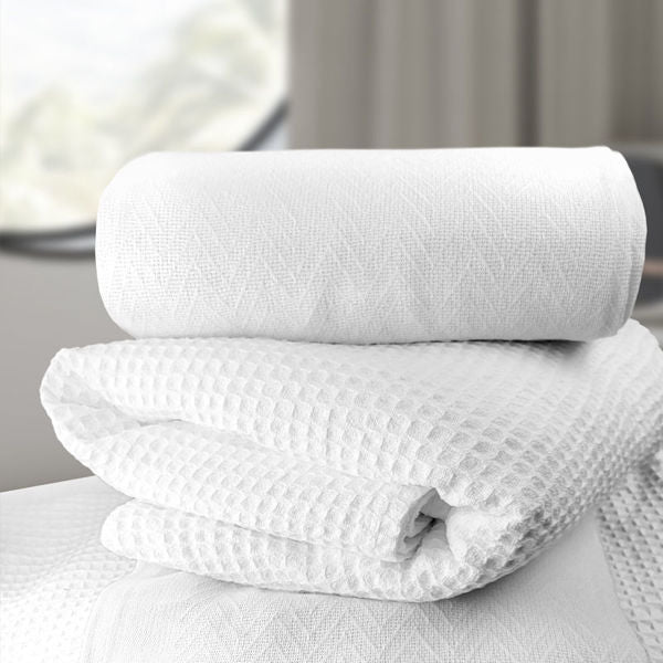 A neatly folded stack of white towels, with a rolled towel on top, sits beside Ganesh Mills Oxford Herringbone Blankets in White/Beige, set against a softly blurred background with light streaming through a window.
