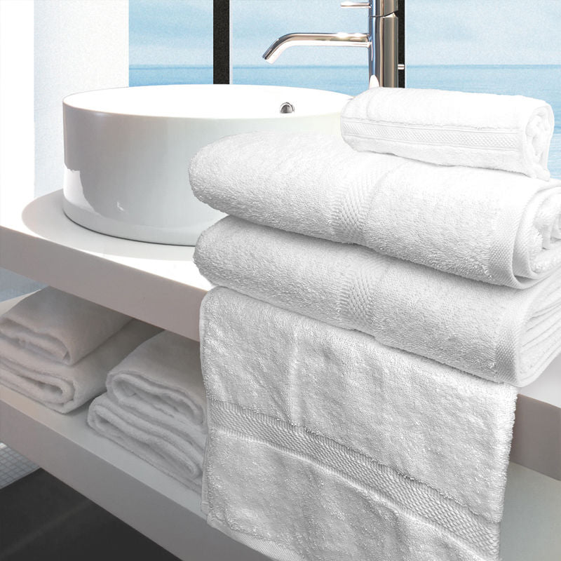 A stack of white Ganesh Mills Oxford Imperiale Bath Towels is folded on a bathroom counter beside a round sink, with more ringspun cotton towels below and a window with a blue view in the background.