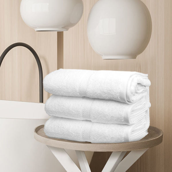Three Ganesh Mills Bath Towel - Double Dobby Oxford Imperiale towels are neatly stacked on a round table in a modern, minimalist room with light wood paneling and two white pendant lights above.