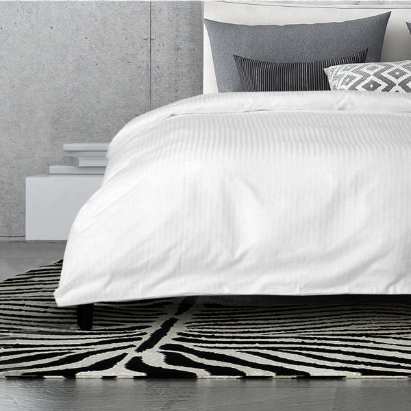 A modern bedroom features a neatly made bed with a Ganesh Mills Bed Topper—5mm Tone On Tone Stripe T250 Mercerized—white comforter, gray and patterned pillows, minimalistic decor, hotel bedding details, and a black-and-white geometric rug.