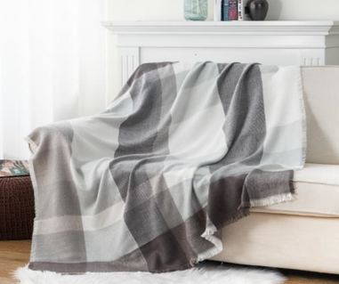 A beige sofa with the Pro Towels Canyon Comfort Throw, featuring soft brown, gray, and cream tones, is draped over the back in a bright room with a white mantle and a fluffy white rug.