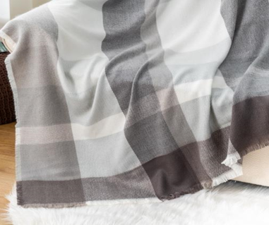 The Canyon Comfort Throw by Pro Towels, featuring soft gray and beige plaid, is draped over a couch with part resting on a fluffy white rug atop a wooden floor.