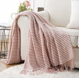 The Pro Towels Charleston Throw, featuring pink and white stripes with fringed edges, is draped over a light-colored sofa to bring cozy charm to the living room.