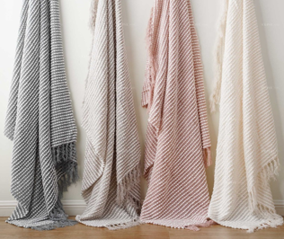 Four Pro Towels Charleston Throw blankets in gray, taupe, blush pink, and cream hang side by side on a wall, their premium textures softly draping down to the wooden floor.