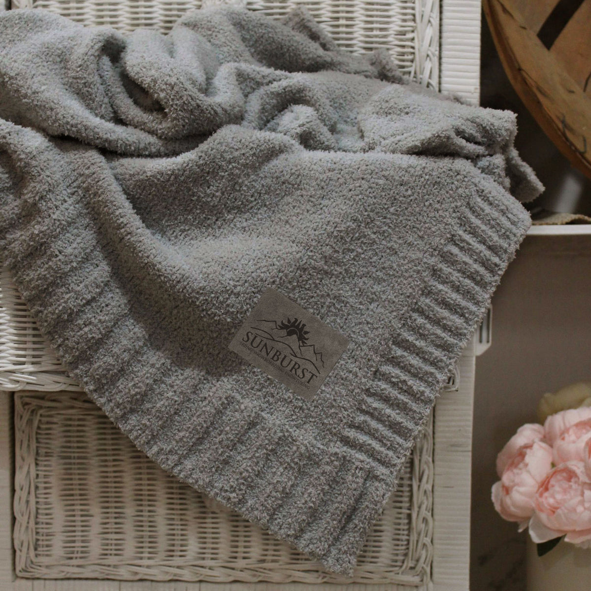A Cloud Nine Soft Throw by Pro Towels in plush gray microfiber drapes over a white wicker chair. A pink peony flower rests in the lower right, with a wooden basket just behind the chair.