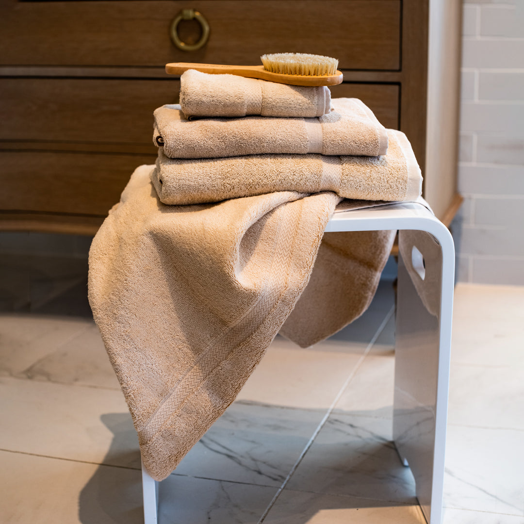 A neatly folded stack of Thomaston Mills Royal Suite Beige Dobby Towels sits on a white stool, topped with a wooden bath brush, in a tiled bathroom with a wooden cabinet in the background.