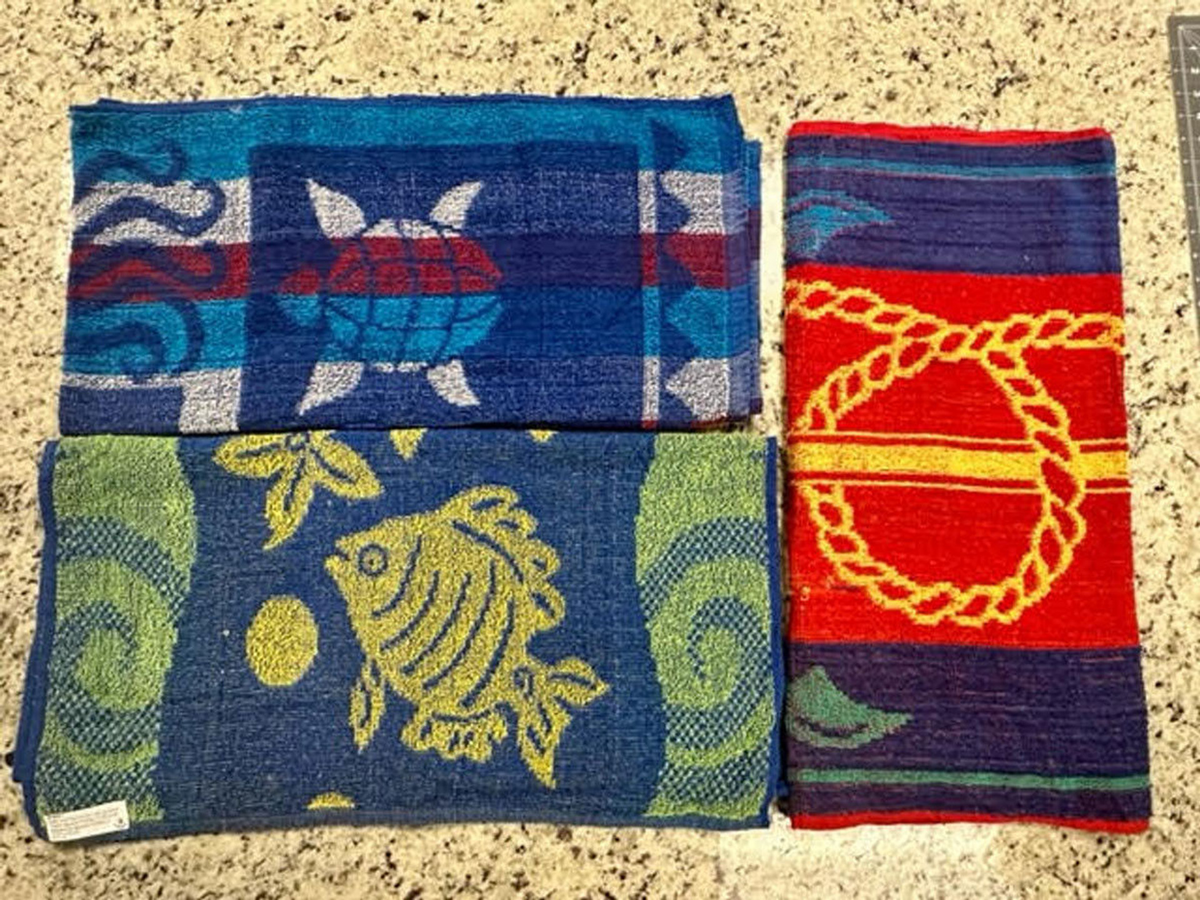 Three Ganesh Mills Promotional Beach Towels—assorted designs including a turtle, fish, and rope loop—are displayed on a speckled countertop. Each cotton jacquard towel features vibrant blues, reds, yellows, and greens for beach style.