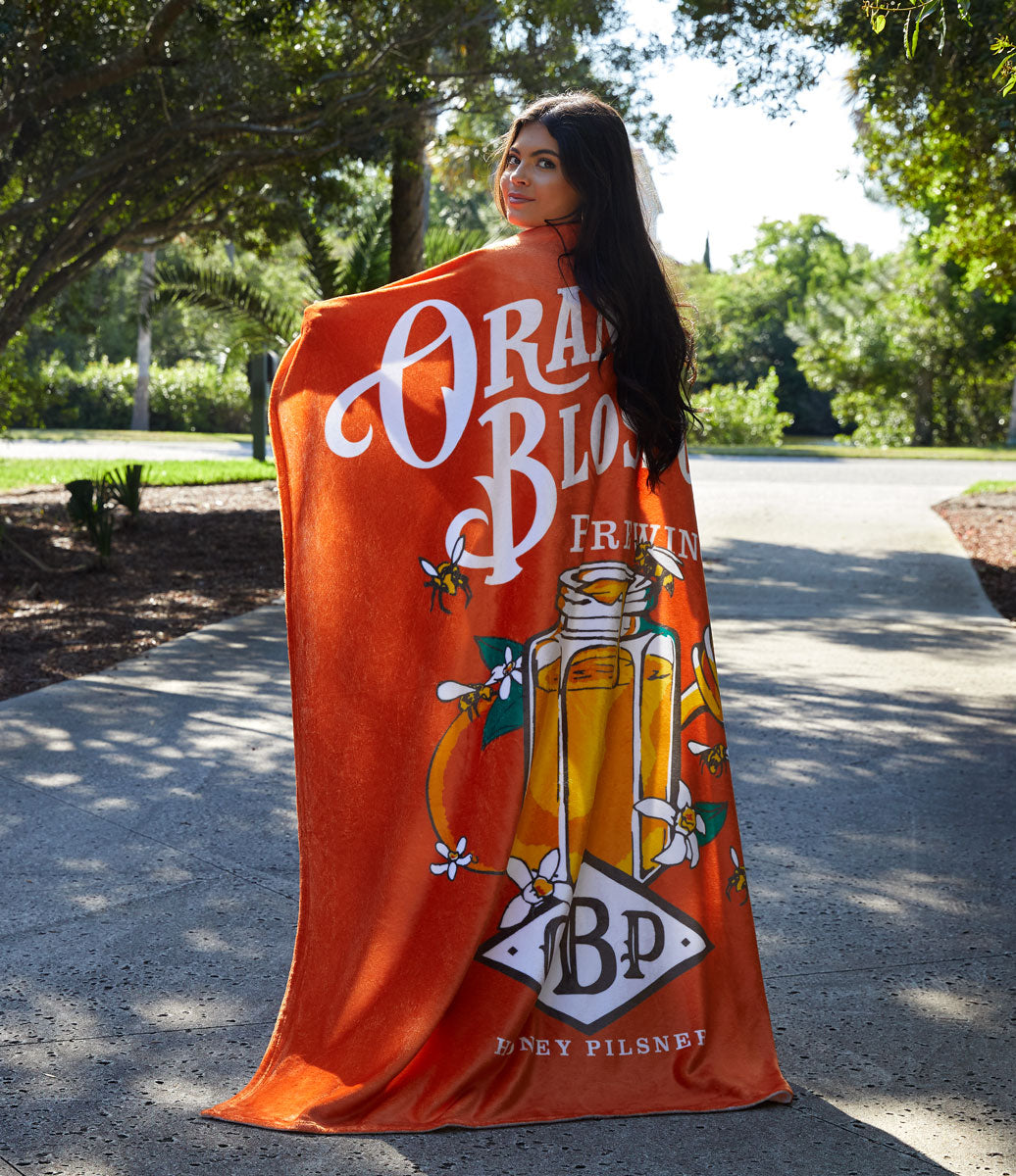 A woman with long dark hair stands on a sunlit path, wrapped in a Pro Vision Plush Throw by Pro Towels featuring "Orange Blossom", a honey pilsner bottle, and white flowers, with trees and greenery in the background.