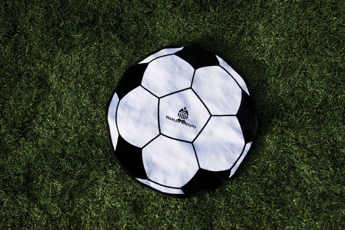 The Pro Towels Soccer Ball Shaped Sport Towel – Fiber Reactive Design lies on green grass, featuring a logo and “HARBOR KNIGHT” in the center—ideal as a team gift or sport towel for any soccer fan.