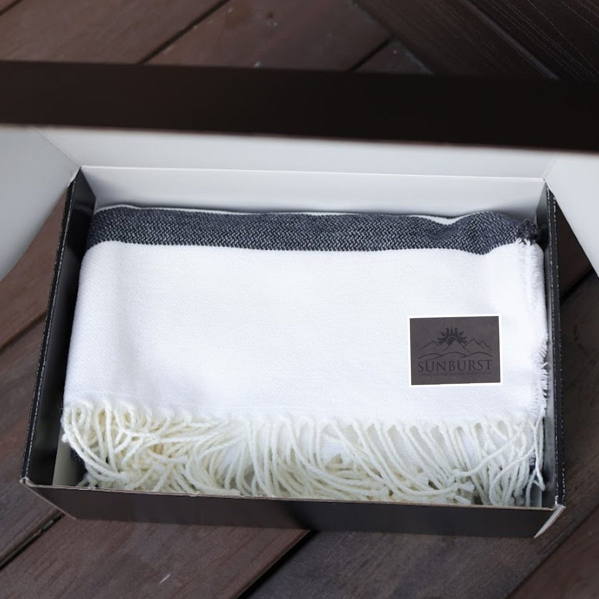 A Four Seasons Striped Blanket by Pro Towels, white with black stripes and fringed edges, sits folded in an open black box on a wooden surface. A tag with the SUNBURST logo is attached to the blanket.