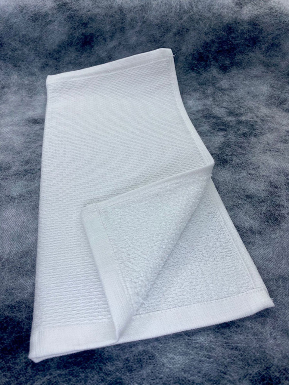 The Ganesh Mills Terry Cloth - White Specialty / Tea Towel is neatly folded on a textured gray surface, revealing its soft, absorbent side and smooth edge. Ideal for use as fingertip towels or enhancing wholesale tea towel collections.