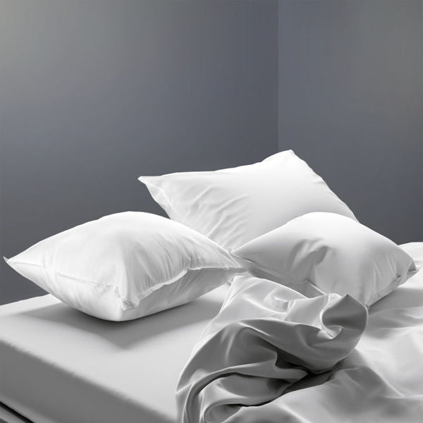 A minimalist bedroom features a neatly made bed with white sheets and three Ganesh Mills Oxford Pillows with Microgel, set against a plain gray wall. The slightly rumpled bedsheet adds a cozy, lived-in touch.