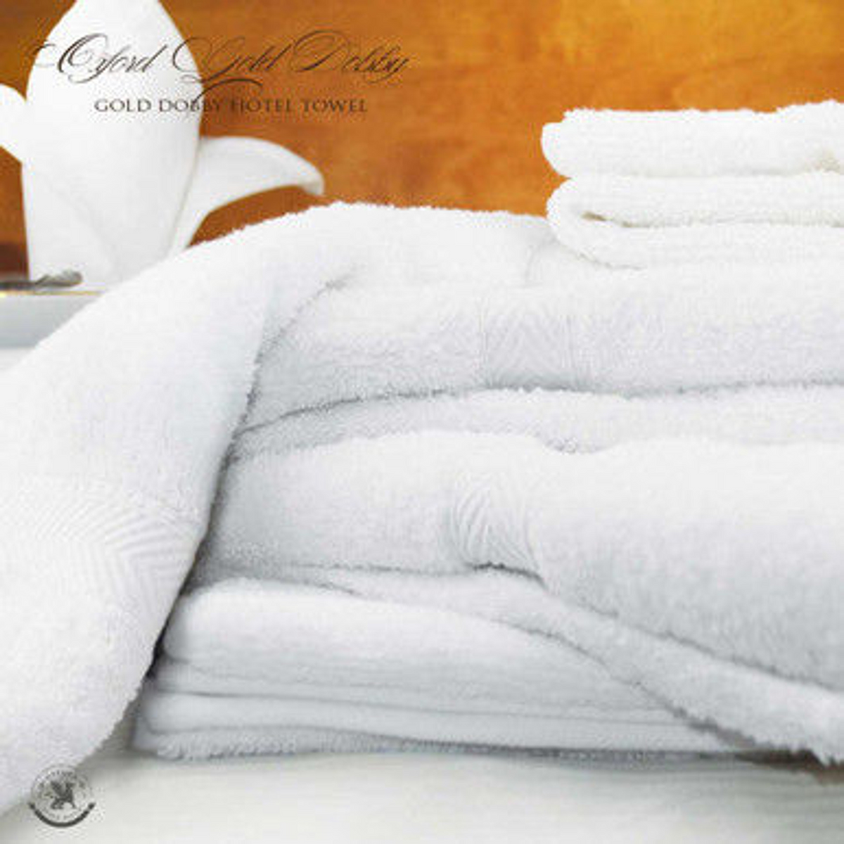 A stack of Ganesh Mills Bath Sheet / Pool Towel - White Oxford Gold Dobby Towels is neatly folded on a surface, with a decorative tissue holder in the background set against a warm, golden wall.