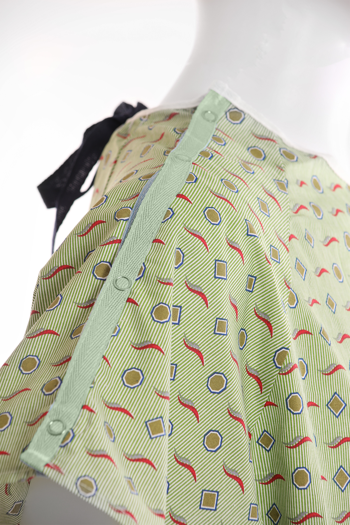 Close-up of a KSE Polyester I.V. Gown with green patterned fabric (squares, circles, red wavy lines), side snap buttons, and black back tie. Shown on mannequin torso. Ideal for bulk orders of medical gowns.