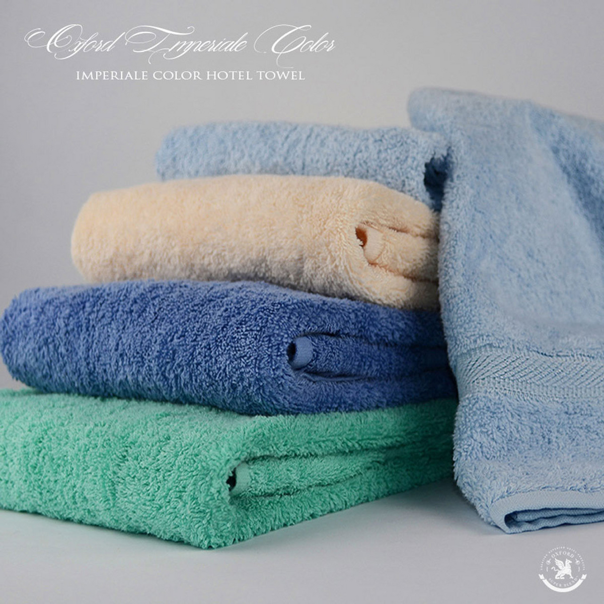 A stack of four pastel blue, yellow, green, and dark blue towels is shown on a light gray background. At the top: "Ganesh Mills Bathmat - Oxford Imperiale Color Towel" with ringspun cotton bathmat options available.