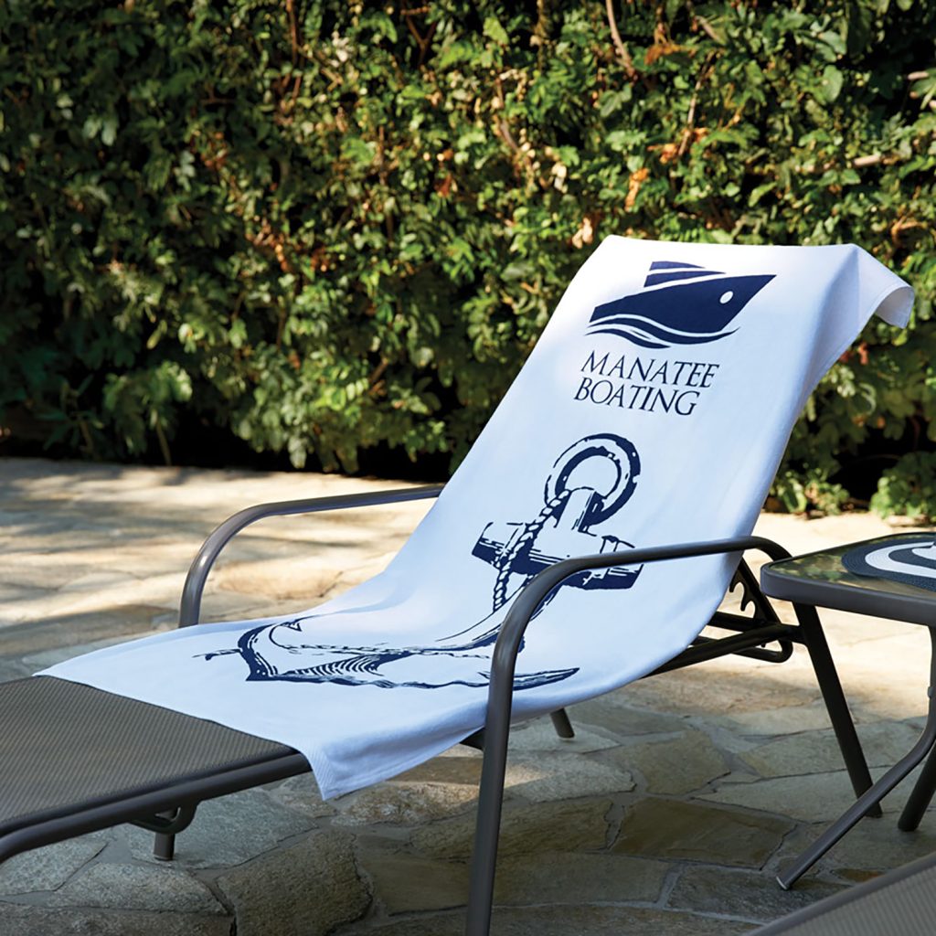 The Pro Towels Beach Towel - Jewel Collection, custom embroidered with a blue anchor and MANATEE BOATING text, is displayed draped over an outdoor lounge chair near a table and greenery.
