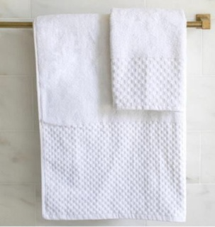 Two white 1Concier Kings Row towels, one large and one small, hang on a brass towel bar against a light tiled wall. Made from a cotton-polyester blend, these towels feature a subtle checkerboard dobby pattern.
