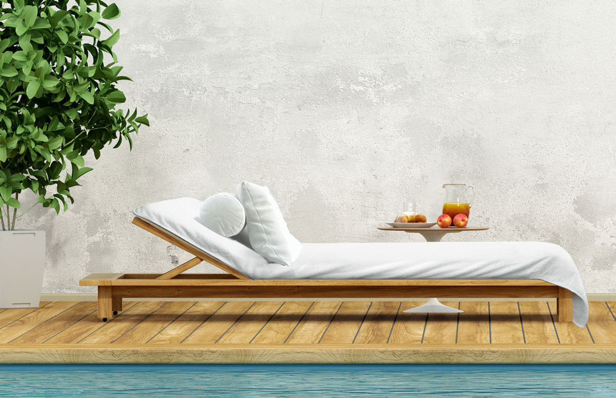 A wooden lounge chair with Ganesh Mills' Oxford Lounge Cover Towel in White/Beige and white cushions sits on a deck by the pool. To the left is a potted plant; a small table with juice and fruit sits against a light wall.