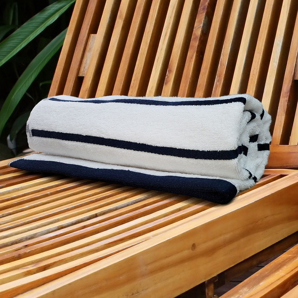 A rolled-up cream-colored Pro Towels Luxury Pool Towel with black stripes rests on a wooden slatted lounge chair, with green leaves in the background.