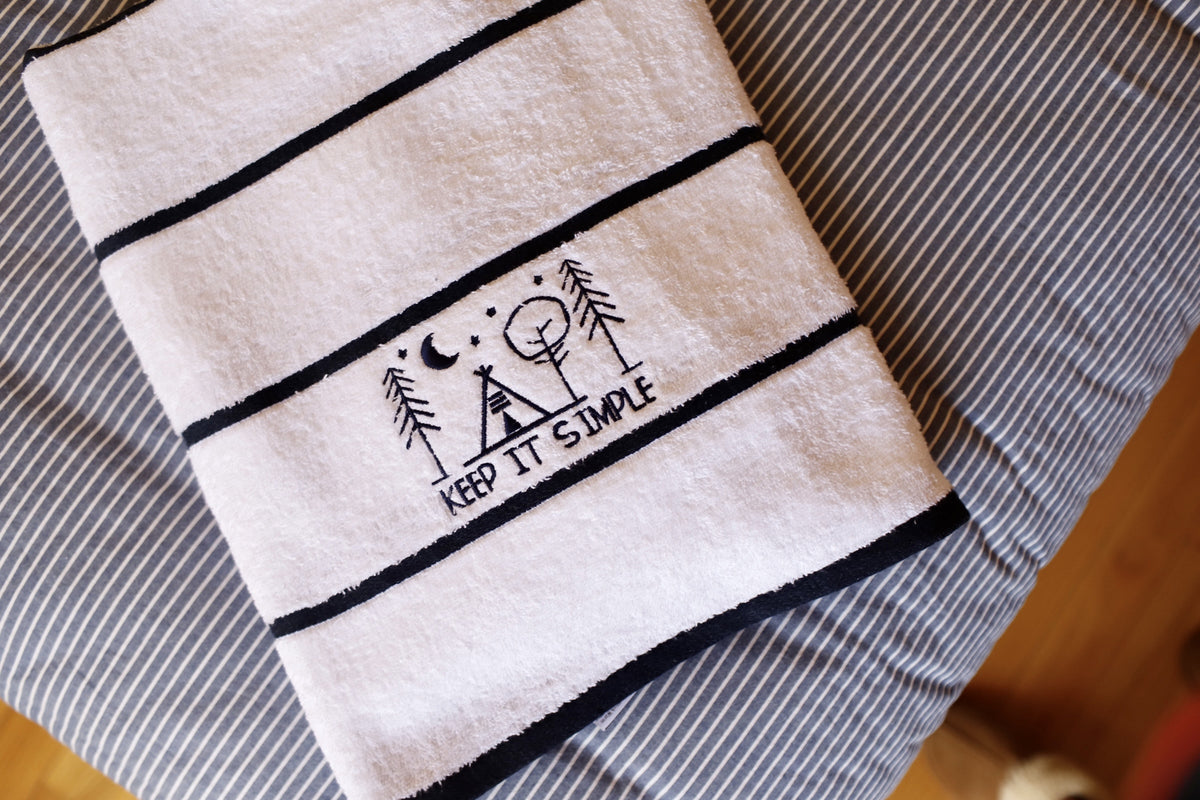 The Pro Towels Luxury Pool Towel features black stripes and custom embroidery of trees, a tent, a crescent moon, and Keep It Simple, displayed on a blue-and-white striped surface.