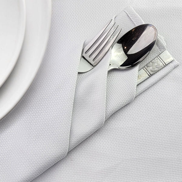 A white Ganesh Mills Napkins - White Momie Banquet Table Linen holds a fork and spoon, neatly folded and placed beside a white plate on a textured white tablecloth.