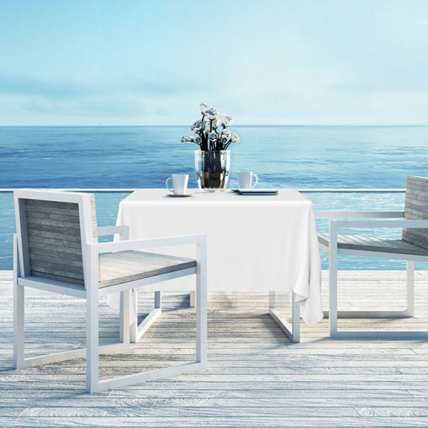 A table with Ganesh Mills’ Oxford Square Table Linen - Multicolor and two modern chairs sits on a deck over a calm blue sea. On the table are hemmed napkins, a metal vase with white flowers, two cups and saucers, and a metal ice bucket.