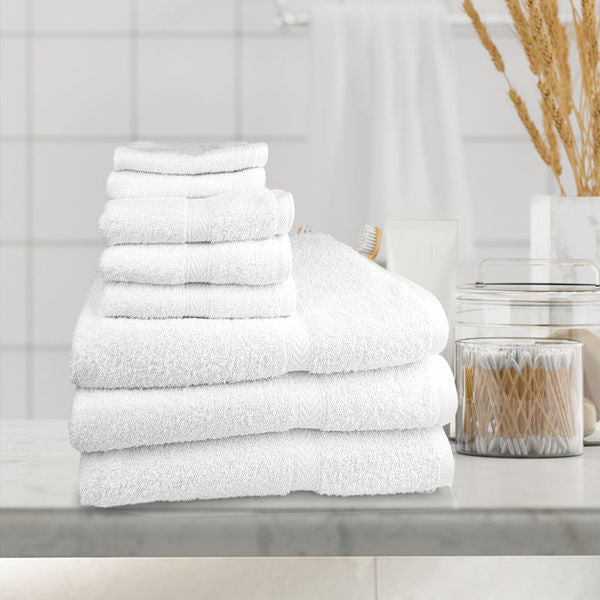 A set of white towels in various sizes is displayed on a marble counter, paired with a plush Ganesh Mills Bathmat - White Oxford Gold Dobby Towel; jars, a brush, and dried reeds add to the elegant backdrop.