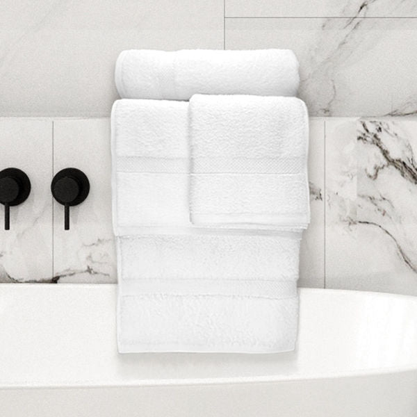 A stack of Ganesh Mills Hand Towel - White Oxford Nuvola Towels, made from ringspun cotton, is neatly arranged on the edge of a white bathtub with light marble tile and black fixtures in the background.