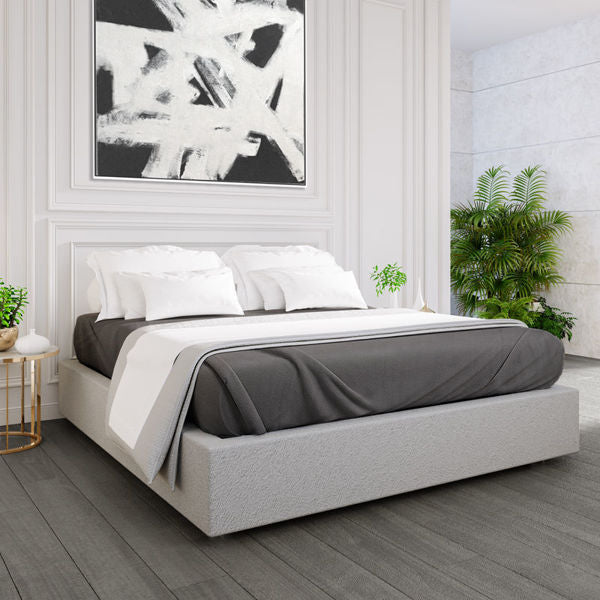 A modern bedroom features a low grey upholstered bed with white bedding and a Ganesh Mills Oxford Polar Fleece Blanket with anti-pilling, double-stitched edges. Green plants, a round side table, and black-and-white abstract art complete the space.