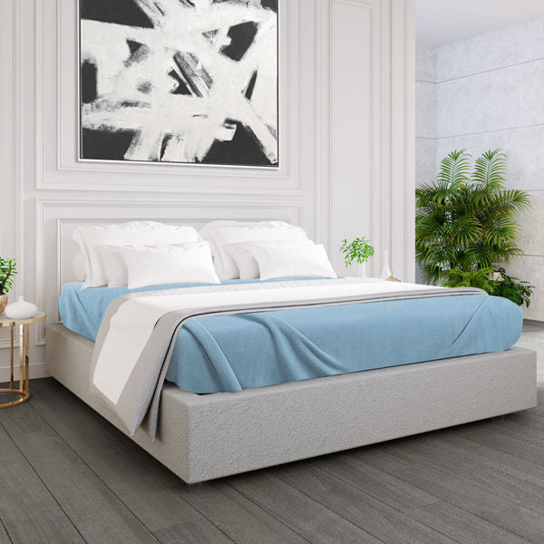 A modern bedroom with a low upholstered bed made up with Ganesh Mills Oxford Polar Fleece Blankets—anti-pilling, double-stitched edges—in blue and white, white walls with molding, abstract black-and-white art, potted plants, and a round bedside table.