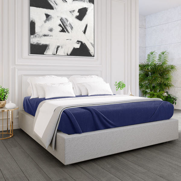 A modern bedroom features a low, light-gray upholstered bed with Ganesh Mills Oxford Polar Fleece Blankets—anti-pilling, double-stitched edges—blue and white bedding, abstract black and white art above the bed, potted plants, white walls, and dark wood floors.